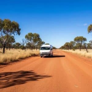 toyota-hiace-bush-tile_1_3c25261e-13c4-4438-a965-5f4282b7d295 - Midland Cylinder Heads White van driving along a straight red dirt road through a dry, sparse savanna with scattered trees under a clear blue sky.